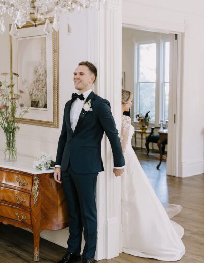 A bride and groom stand back-to-back, holding hands around a corner. The groom smiles while dressed in a tuxedo, and the bride wears a white gown. A chandelier hangs above.