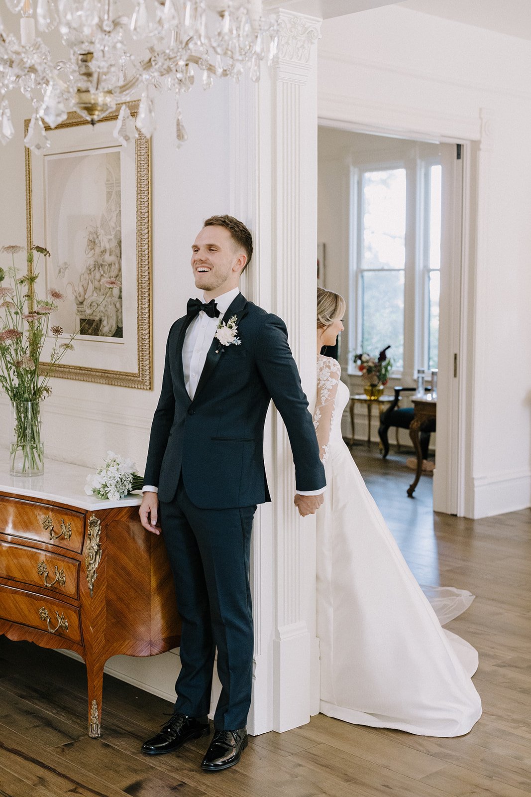 A bride and groom stand back-to-back, holding hands around a corner. The groom smiles while dressed in a tuxedo, and the bride wears a white gown. A chandelier hangs above.