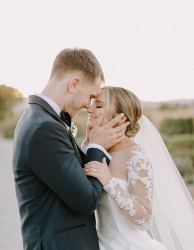 A couple in wedding attire embrace outdoors, smiling gently, with a soft background of greenery and sky.