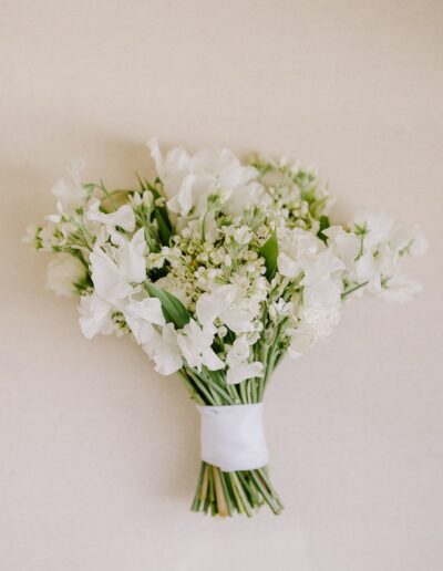 A bouquet of white flowers with green stems and small buds, wrapped with a white ribbon, against a plain beige background.