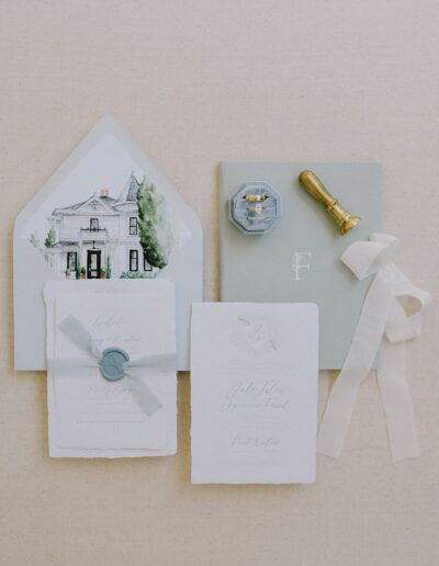 A collection of elegant stationery featuring invitations, an envelope with a house illustration, a wax seal stamp, a ring box, and a ribbon on a beige background.