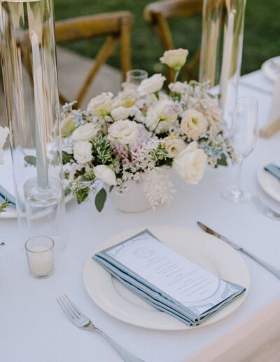 Elegant table setting with white and pastel flowers in a vase, two tall candles, and neatly arranged plates with folded menus on a white tablecloth.