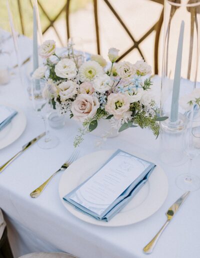 Elegant table setting with plates, blue napkins, menu cards, white flowers, and blue candles on a white tablecloth.