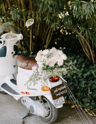 White scooter decorated with a floral arrangement on the seat, a small "Just Married 9-10-2023" sign at the back, parked near green foliage.