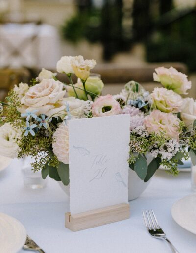 A floral centerpiece with pastel flowers and greenery on a white tablecloth. A card with "Table 20" is placed in a wooden holder. Plates and a fork are visible.