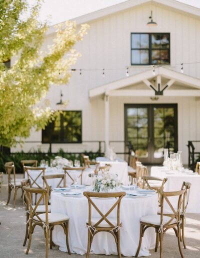 Outdoor wedding reception setup with round tables covered in white tablecloths, wooden chairs, and floral centerpieces in front of a white building.