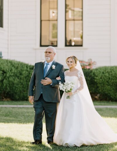 A bride in a white gown and veil walks arm-in-arm with an older man in a suit, holding a bouquet of flowers. They are outside, with greenery and a white building in the background.