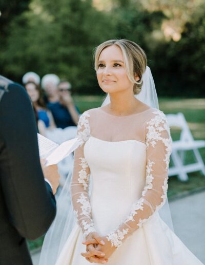 A bride in a white dress with lace sleeves stands outdoors, hands clasped, facing a person holding a paper. White chairs and greenery are in the background.