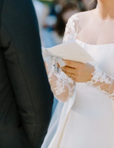 Bride in a white dress reads vows from paper while standing next to a groom in a black suit at an outdoor ceremony.