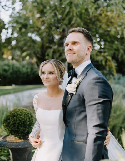 A bride and groom stand outdoors, embracing and looking ahead. They are dressed in formal wedding attire, surrounded by greenery.