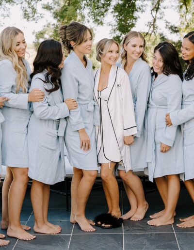 A group of women wearing matching bathrobes stand together on a balcony, smiling. Trees and a railing are visible in the background.