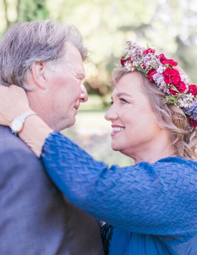 A couple stands outdoors, facing each other closely. The woman with a flower crown smiles at the man, who wears a gray jacket. The background is blurred greenery.