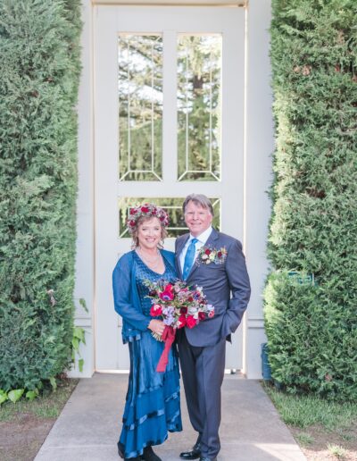 A couple stands in front of a white door flanked by tall greenery. The woman holds a bouquet, and both wear formal attire with floral accents.