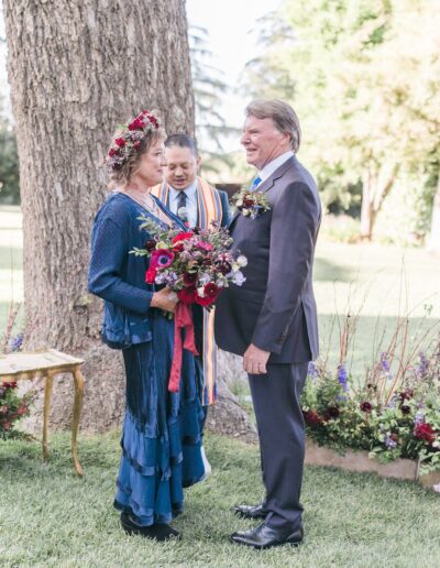 A couple stands together outdoors during a wedding ceremony, with a person officiating. They are surrounded by flowers and greenery.