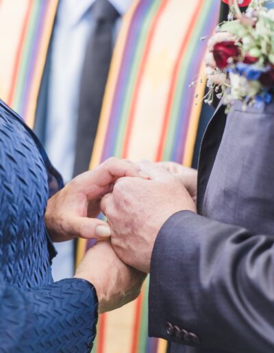 Two people holding hands during a ceremony, with one wearing a dark suit and floral corsage, and the other in a blue textured garment. A person with a colorful stole stands nearby.