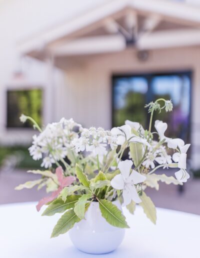 A white vase with white flowers and green leaves on a table, set outdoors near a modern house.