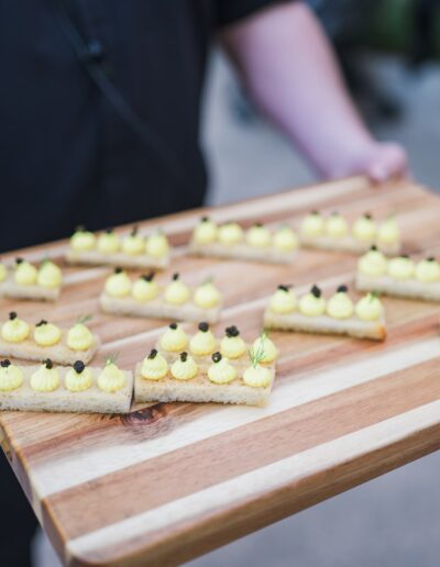 Person holding a wooden tray with multiple rectangular appetizers topped with yellow cream and small garnishes.