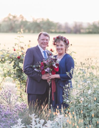 A couple stands outdoors, smiling and holding a bouquet. The man is in a suit, and the woman wears a blue dress with a floral crown. They are surrounded by greenery and flowers.