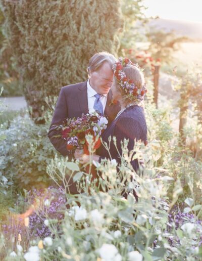 A couple embraces in a garden during sunset, surrounded by lush greenery and flowers. The woman wears a floral wreath.