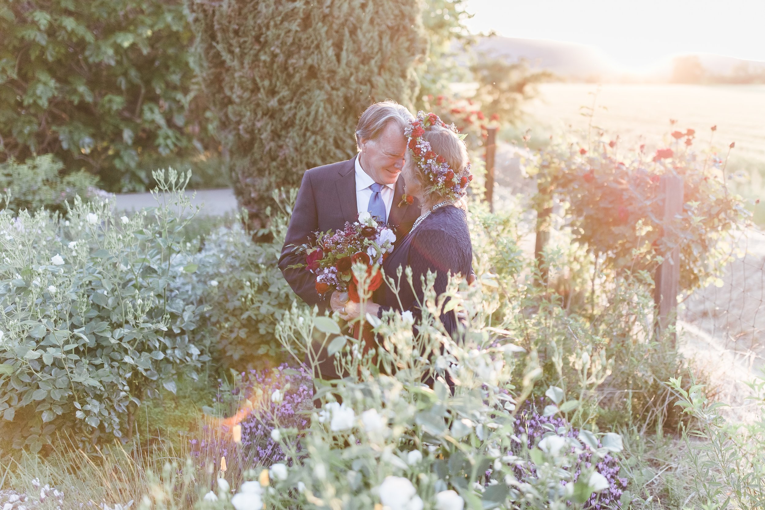 A couple embraces in a garden during sunset, surrounded by lush greenery and flowers. The woman wears a floral wreath.