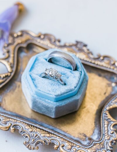 A diamond ring in a light blue velvet box on an ornate metal tray with a blue flower and a yellow-centered white flower nearby.