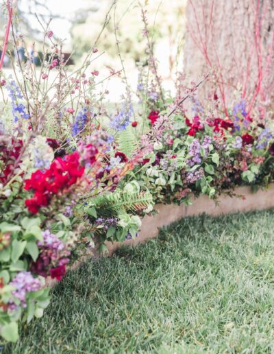 A floral arrangement with red, purple, and green flowers is placed beside a tree. A small gold table stands on green grass nearby.