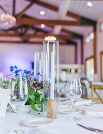 Elegant table setting with a tall glass-enclosed candle, wine glasses, and purple flower centerpiece, under wooden beams in a softly lit room.