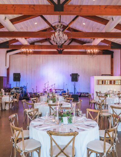 Spacious event hall with round tables set for dining, floral centerpieces, a wooden ceiling with beams, chandeliers, and a stage with musical instruments.