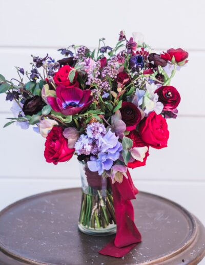 A bouquet of red, pink, and purple flowers with greenery in a glass jar, placed on a round brown table against a white background.