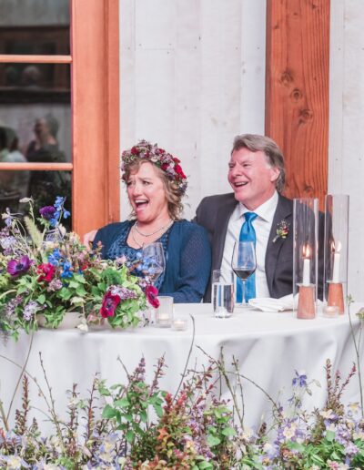 A couple sits at a decorated table, smiling and laughing. The table is adorned with flowers, and candles are lit. They are indoors, near large windows.