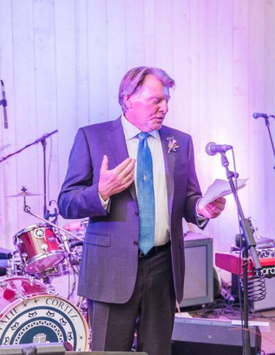 A man in a suit speaks into a microphone, holding a paper. Musical instruments, including a drum set and keyboard, are in the background.