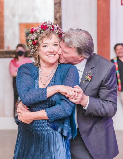 An older couple embraces and smiles; the man kisses the woman's cheek. She wears a floral crown and blue dress, and he is in a suit with a boutonniere. People and a mirror are in the background.