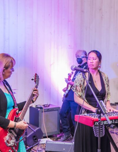 A band performing on stage with guitars, keyboard, and drums. Musicians are wearing casual clothing, and one is wearing a mask. The stage is lit with pink and purple lights.