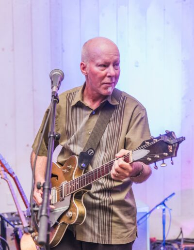 Man playing an electric guitar on stage with a microphone in front, wearing a brown shirt.