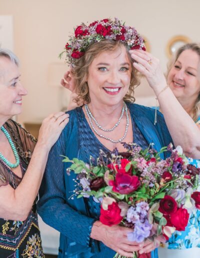 Three women standing together in a bedroom; two women adjust the floral headpiece of the smiling woman holding a bouquet in the center.