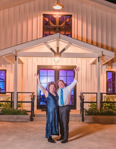 A couple stands with raised arms in front of a brightly lit barn-style venue at night, surrounded by tables and plants.