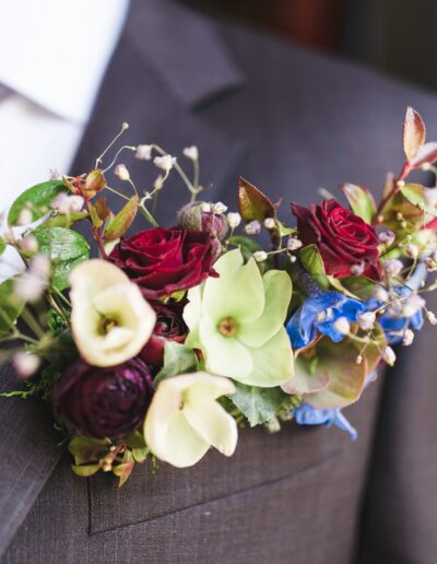 A close-up of a man's suit with a vibrant floral boutonniere featuring red, green, and blue flowers tucked in the lapel pocket.