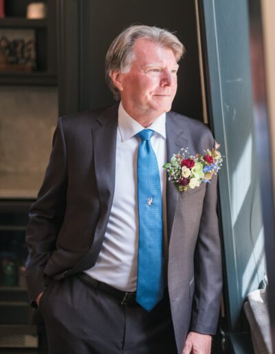 A man in a suit with a blue tie and a floral boutonniere stands by a window, looking outside.