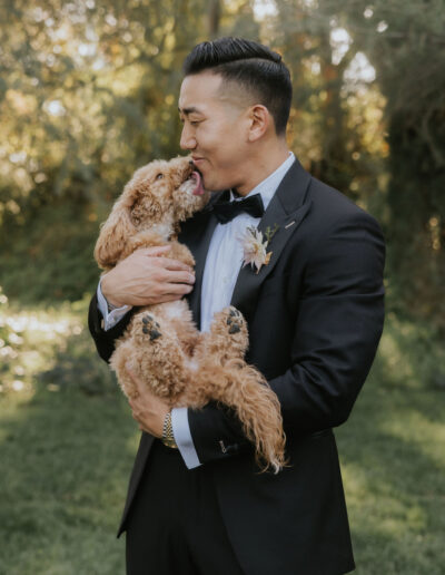 A man in a suit holds a small dog in a grassy area. The dog licks the man's face affectionately.