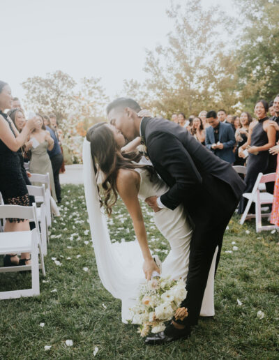 A bride and groom kiss while the groom dips the bride after their outdoor wedding ceremony, surrounded by guests who are clapping.