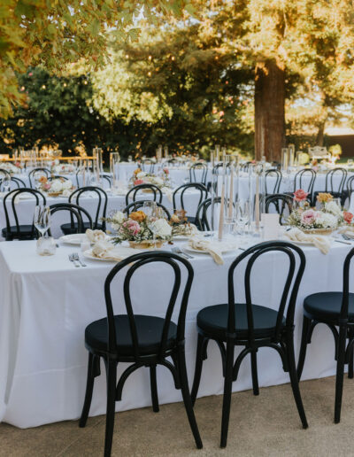 Outdoor dining setup with white tablecloths, black chairs, floral centerpieces, and candles under trees with sunlight filtering through leaves.