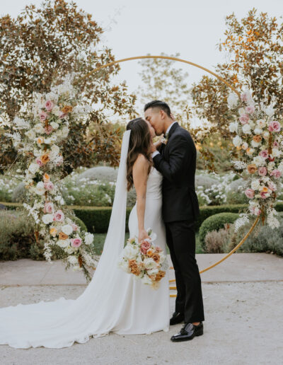 A bride and groom share a kiss in front of a circular floral arch. The bride holds a bouquet, and both are dressed in wedding attire.