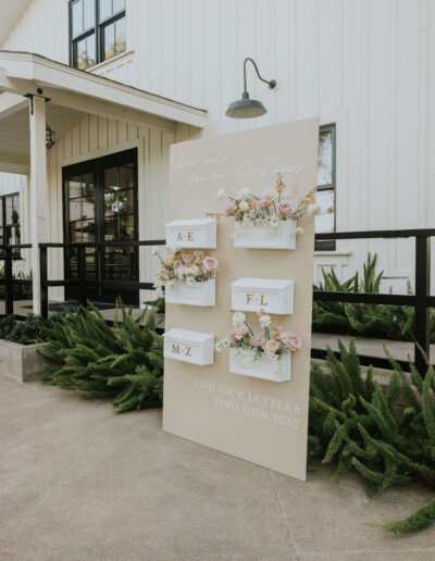 Wedding seating arrangement board with floral decorations, displaying guest letters A-E, F-L, and M-Z in white boxes. The board is in front of a white building with large windows.