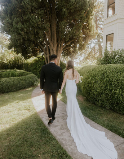 A couple in formal attire walks hand in hand on a garden path, next to green hedges and trees.