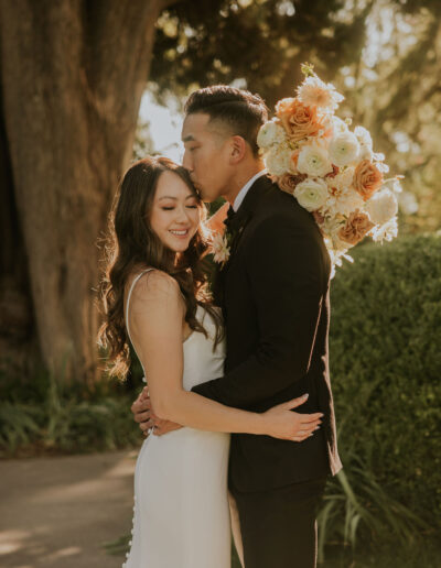 A couple embraces outdoors. The man kisses the woman's forehead while holding a bouquet of roses. They both appear happy.