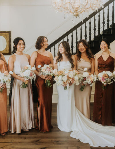 A bride stands with six bridesmaids in elegant dresses, holding bouquets, in a room with a chandelier and staircase.