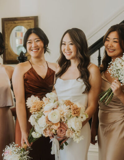 A bride in a white dress stands with five bridesmaids in various shades of brown and blush dresses, holding bouquets of flowers, in a well-lit room near a staircase.