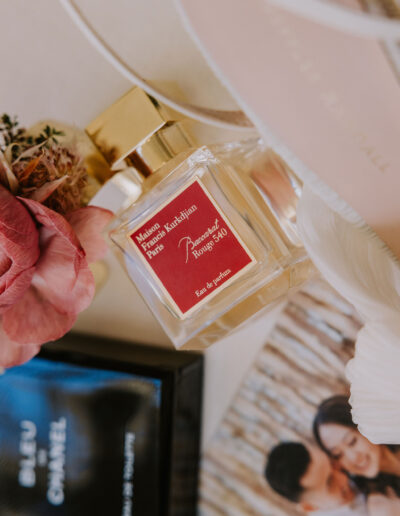 A bottle of Maison Francis Kurkdjian Baccarat Rouge 540 perfume is placed on a flat surface with pink flowers, a black Chanel box, and a photo in the background.