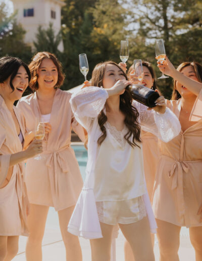 A group of women in matching robes celebrate by a pool, one drinking from a champagne bottle while others hold glasses, smiling and laughing in the sunlight.