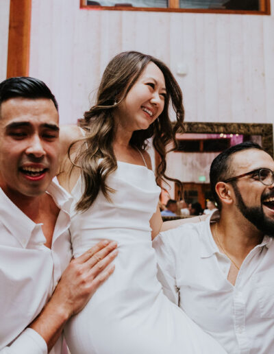 A woman in a white dress is lifted by two men in a festive indoor setting. Everyone appears to be having fun.